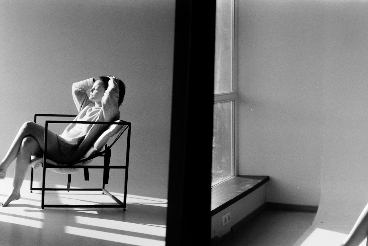 Black and white portrait of a woman relaxing on a chair in a minimalist room with window light.